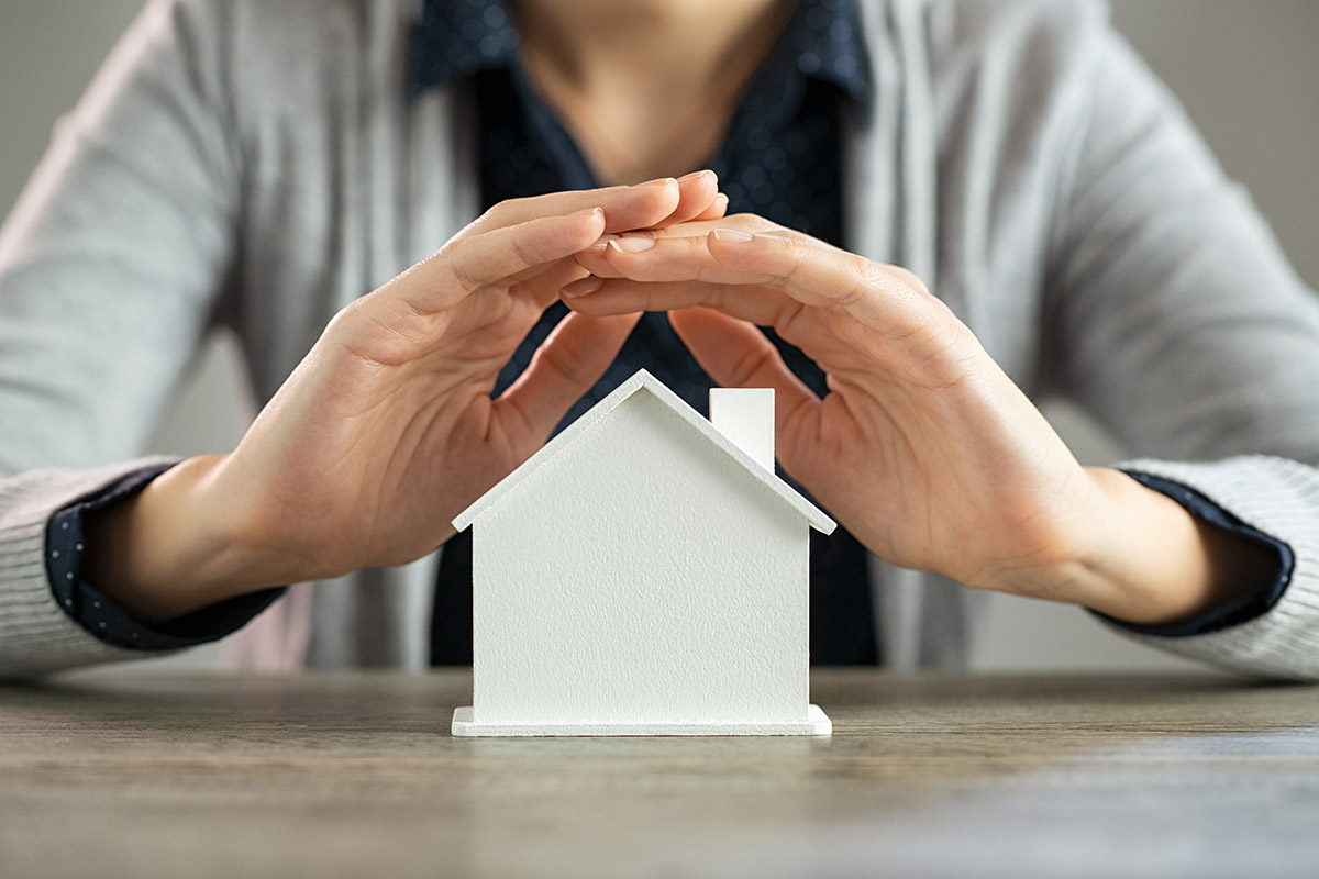 Close up of young woman hands protecting a model of house on table. Detail of woman holding hands over cardboard figure of house at desk. Woman protecting her real estate investment on scale model.